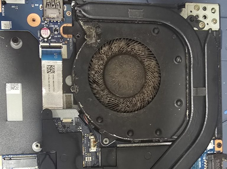 The inside of a gaming laptop opened on a repair bench, with the cooling fan blades completely covered by a thick layer of grey dust